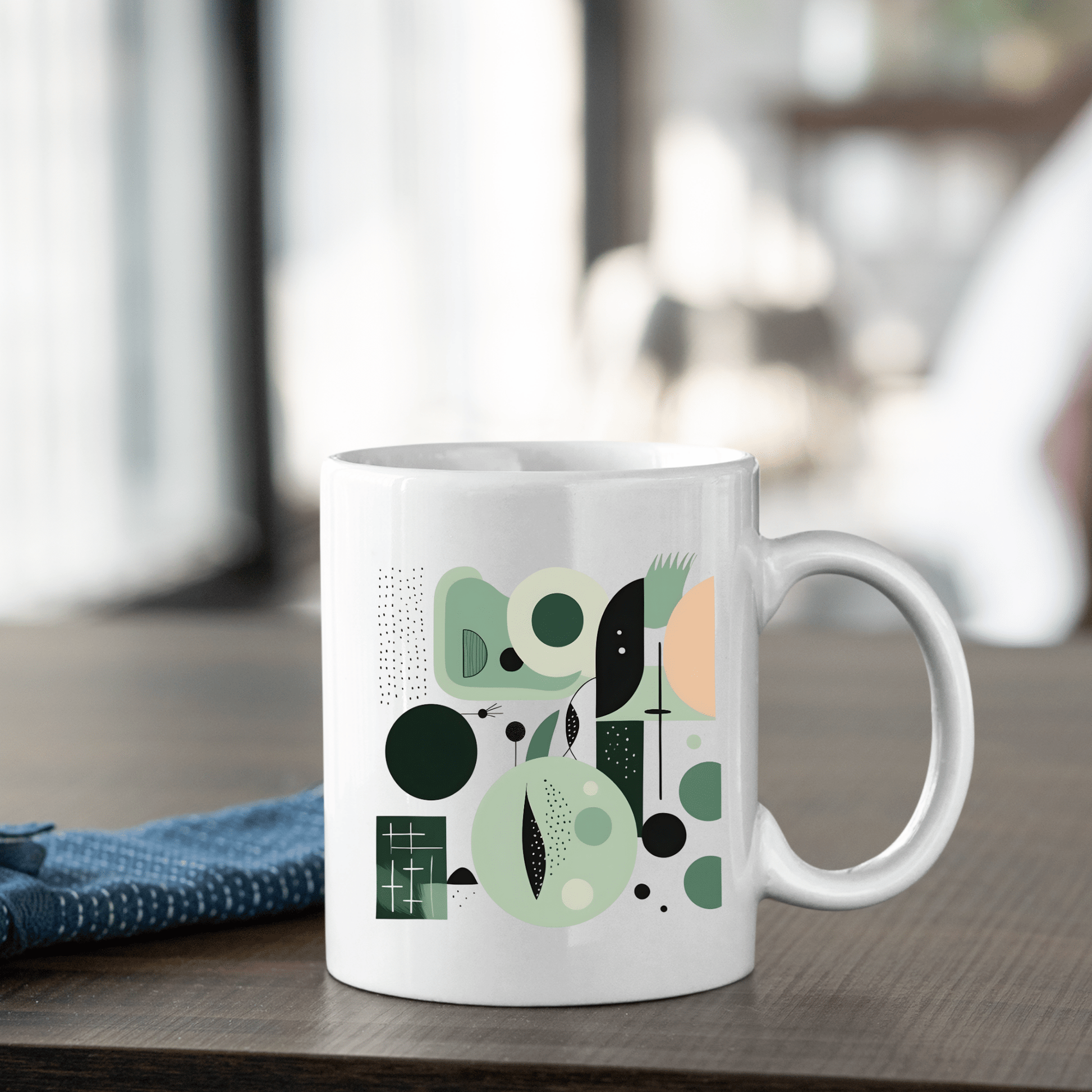 The Spring Geometry Mid Century Modern Mug, crafted from white ceramic and adorned with a green and black abstract design, sits on a wooden table beside a folded blue cloth against softly blurred nature-inspired décor.