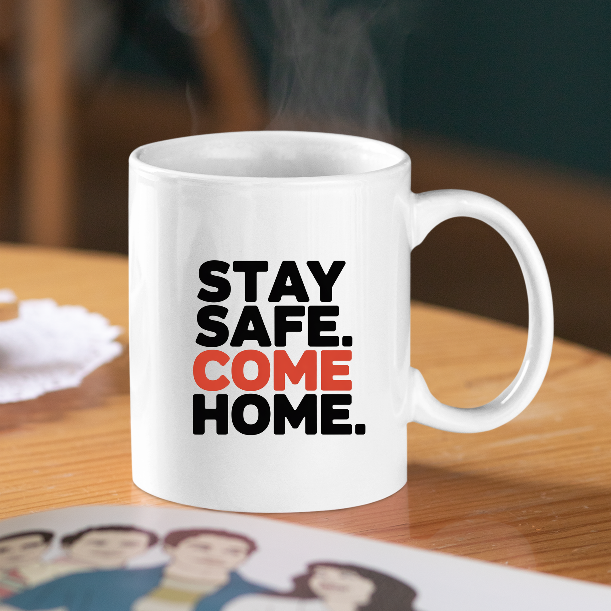 A Stay Safe Come Home Mug | Gift for First Responders, featuring the words “STAY SAFE. COME HOME.” in bold black and orange letters, sits on a wooden table. Steam rises from this white mug designed for police officers, while a partially visible photo with illustrated people adds a personal touch—making it a thoughtful gift for first responders.
