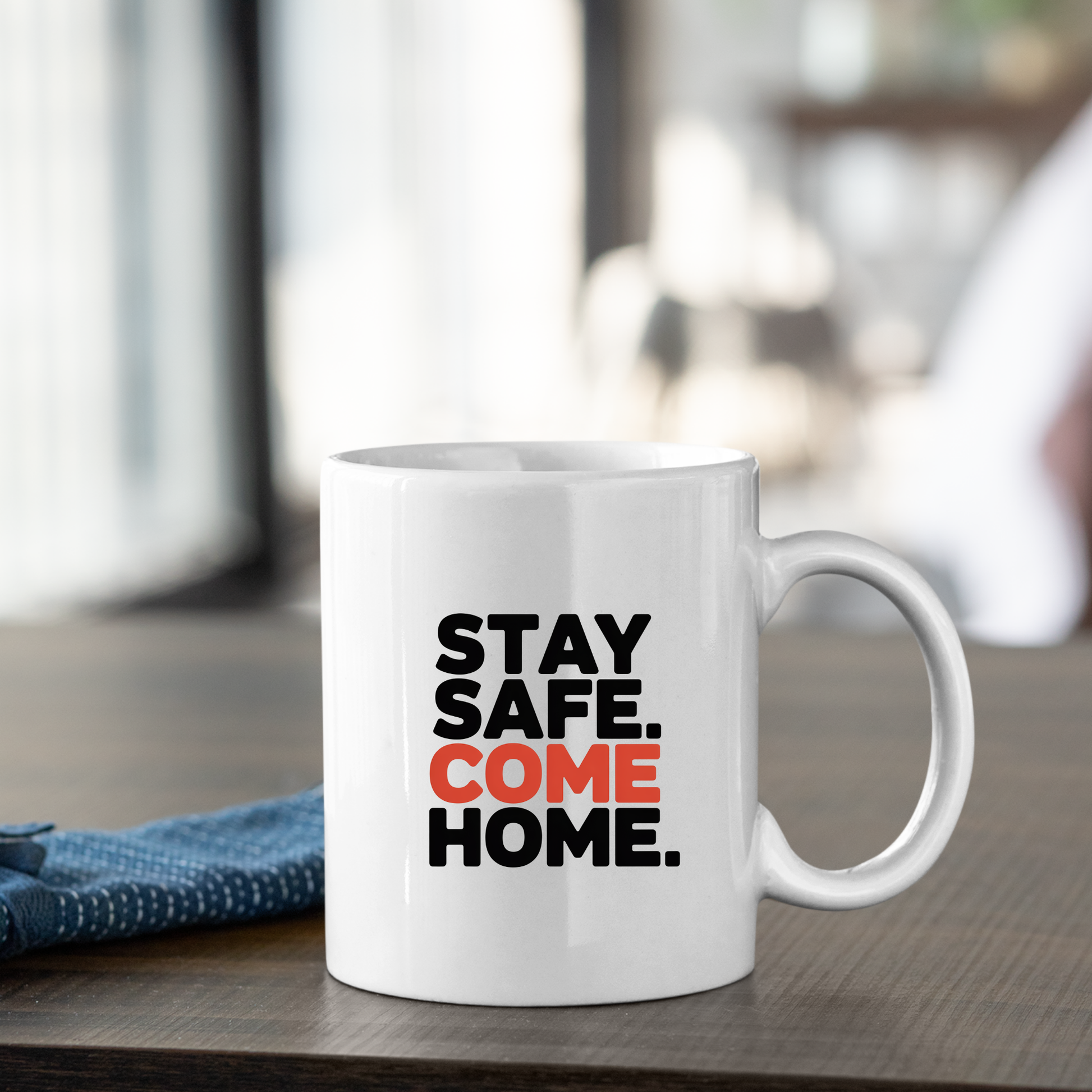 The Stay Safe Come Home Mug | Gift for First Responders rests on a table, featuring the message "STAY SAFE. COME HOME." in bold black and orange lettering. Ideal as a thoughtful police officer mug or first responder gift, it is paired with a blue cloth and set against a softly blurred background.