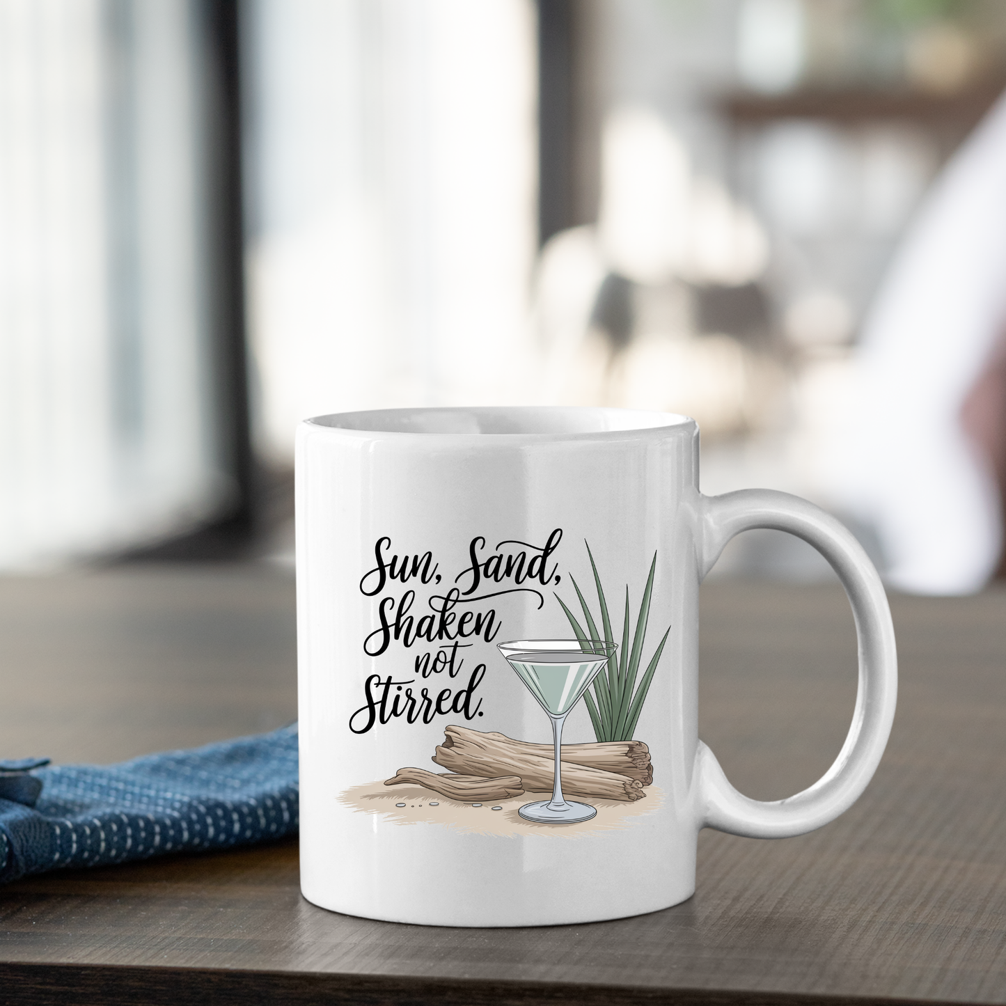 The Sun Sand Shaken Not Stirred Mug | Beach Lover Gift is a white, humorous vacation mug featuring a graphic of a martini glass, driftwood, and greenery. The mug displays the text "Sun, Sand, Shaken not Stirred," making it perfect as a coastal gift or beach coffee mug. It is photographed on a wooden table beside a folded blue cloth.