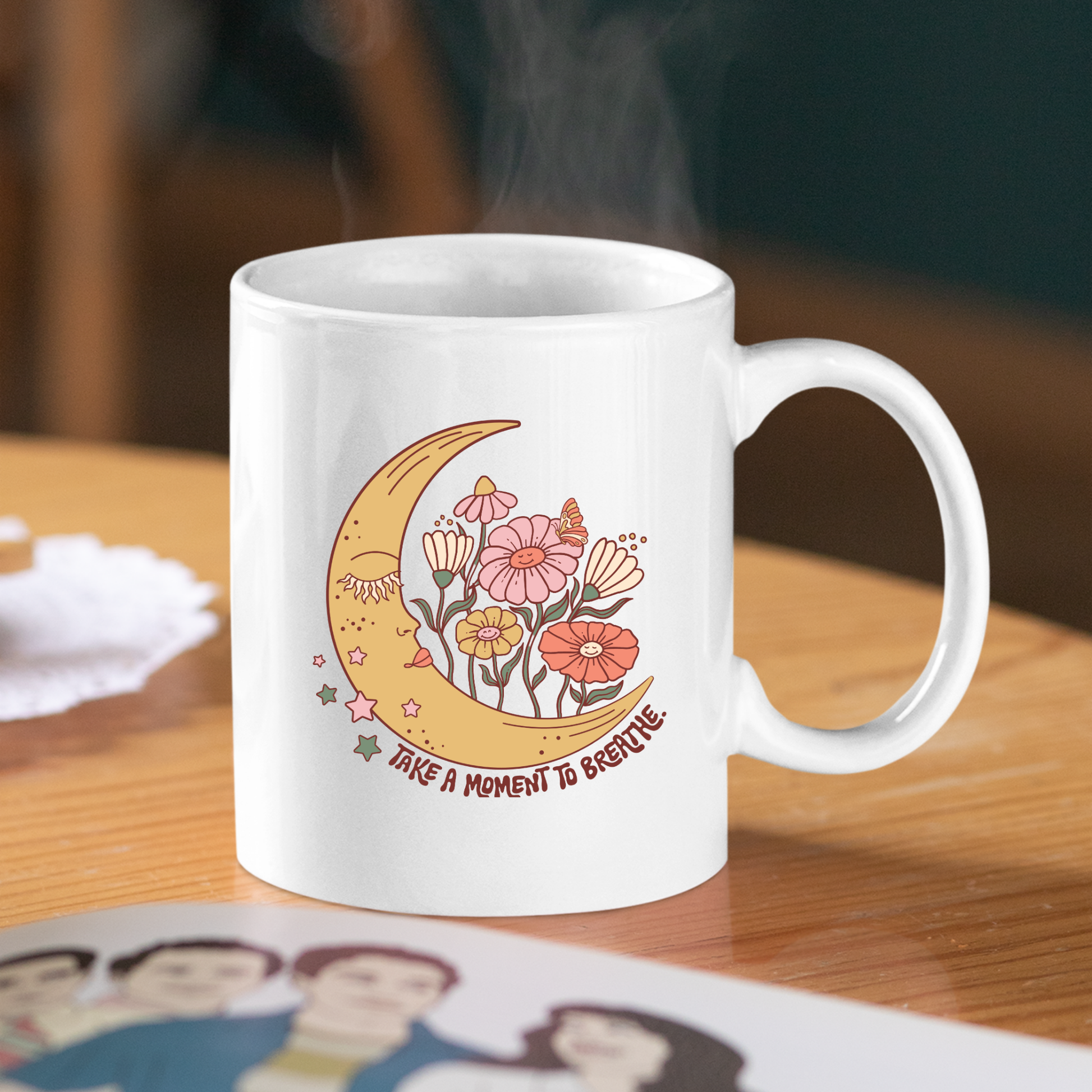 The Take a Moment to Breathe Mug | Mindfulness Coffee Mug features a white design with a crescent moon, colorful flowers, and the text “Take a moment to breathe.” Set on a wooden table with steam rising from it, this inspirational coffee mug makes the perfect meditation gift.