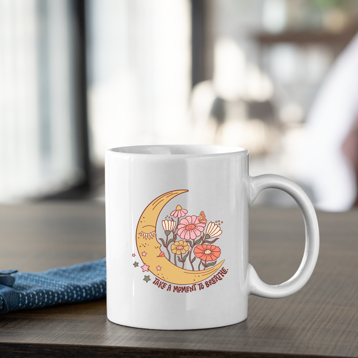 The Take a Moment to Breathe Mug | Mindfulness Coffee Mug rests on a table, its pristine white surface featuring an elegant crescent moon, delicate flowers, and shining stars. Beneath the artwork, the phrase "Take a moment to breathe" is gracefully printed, making this mug an ideal choice for meditation or as a thoughtful self care gift. Beside the mug lies a neatly folded blue napkin.